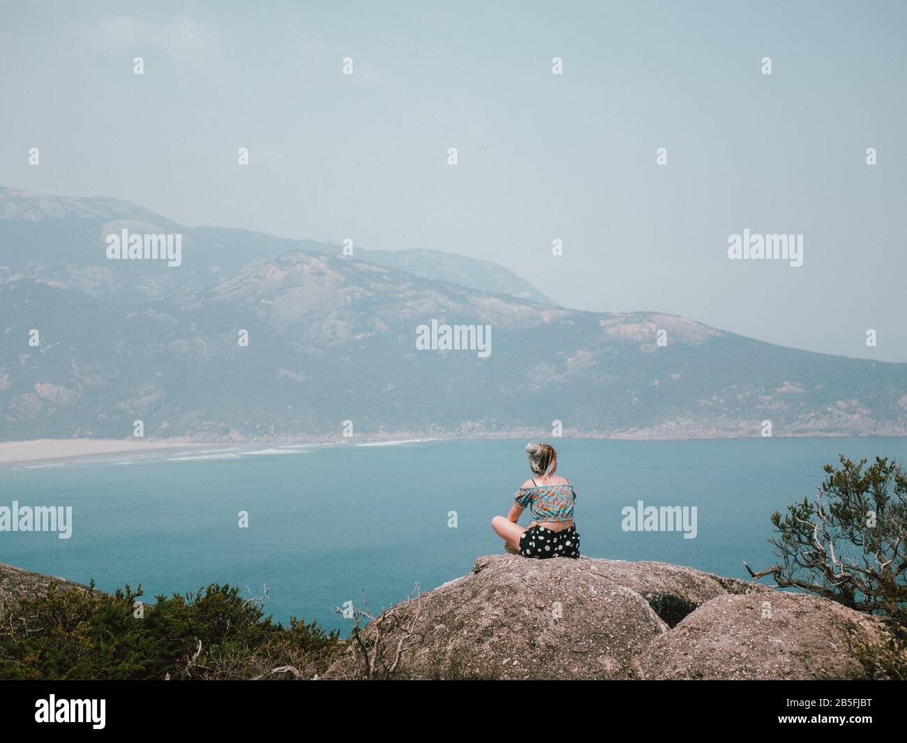 A young woman sitting on the edge of a rock at the top of a mountain looking over a beautifully blue ocean Stock Photo