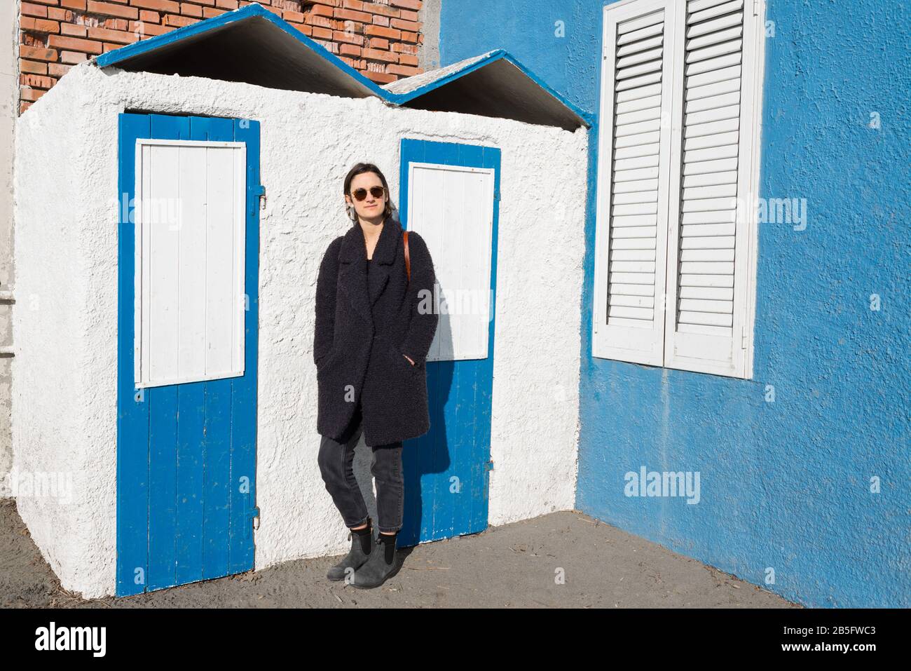 Woman model wearing sunglasses and winter coat in front of seaside cabins painted in vintage pastel blue and white Stock Photo