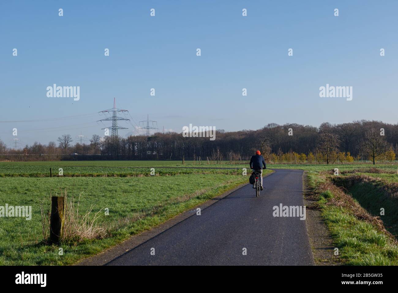 Outdoor sunny view of cyclist ride a bicycle on small road in countryside area with agricultural field and meadow in winter season against blue sky. Stock Photo