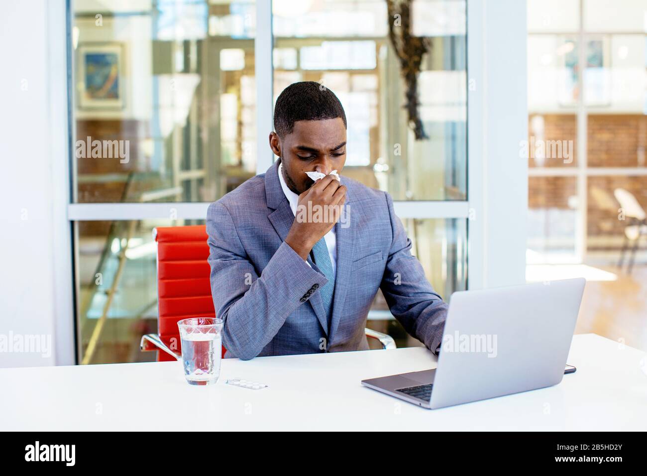 Portrait of a sick young man in business suit having a cold,  blowing his nose while sitting behind desk at work with computer Stock Photo