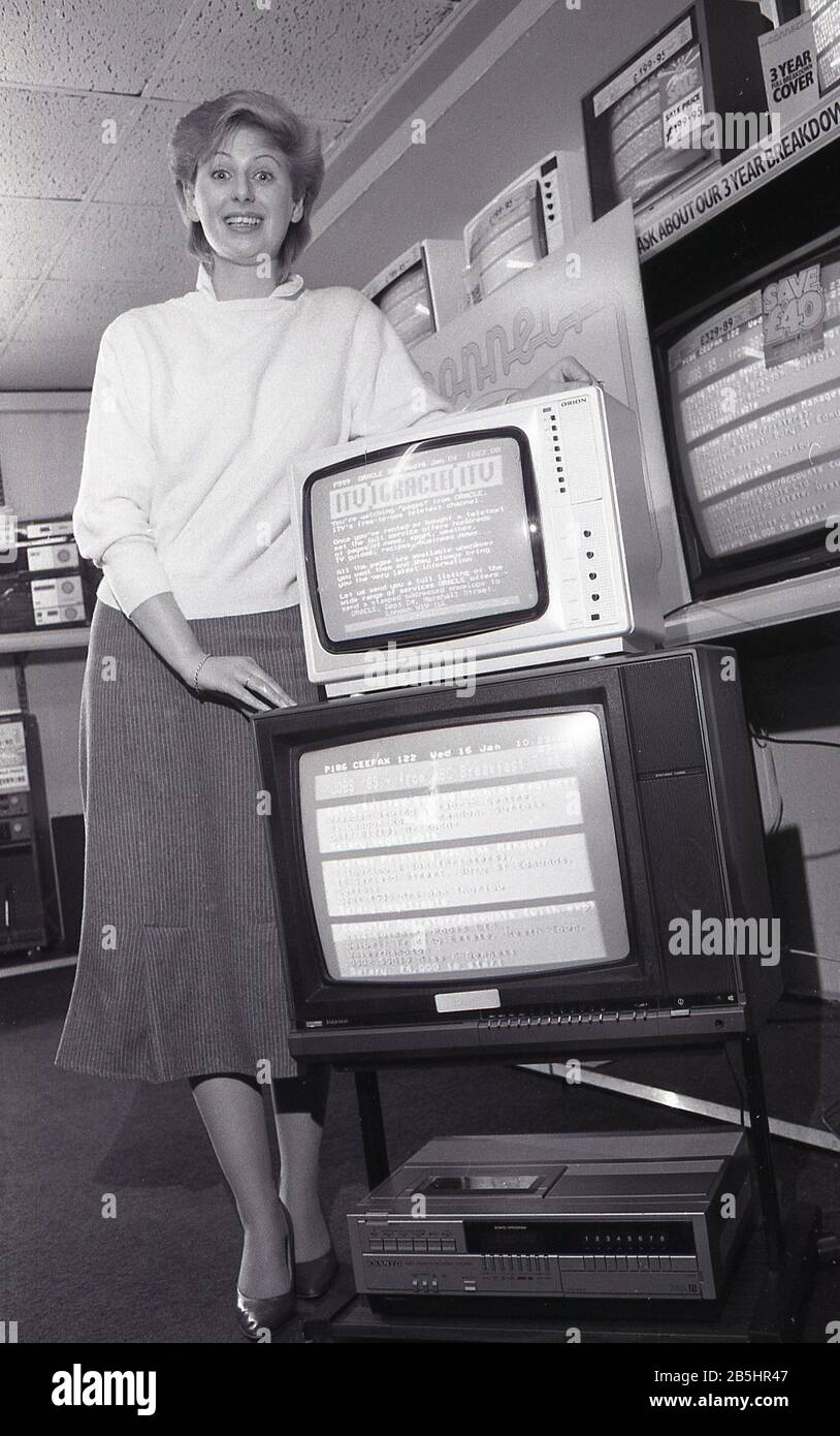 1980s, historical, a saleswoman standing by a display in a retail store of the latest televisions and a VHS player, England, UK. On one of the screens is the ITV teletex. Stock Photo