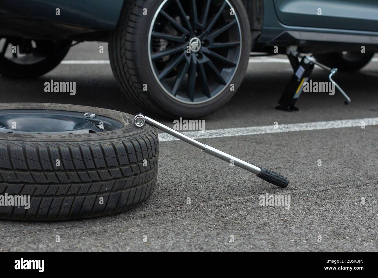 A flat tire car wheel and screwdriver are on a asphalt road on the broken Skoda Rapid background. Jack is lifting up a vehicle. Automobile service Stock Photo