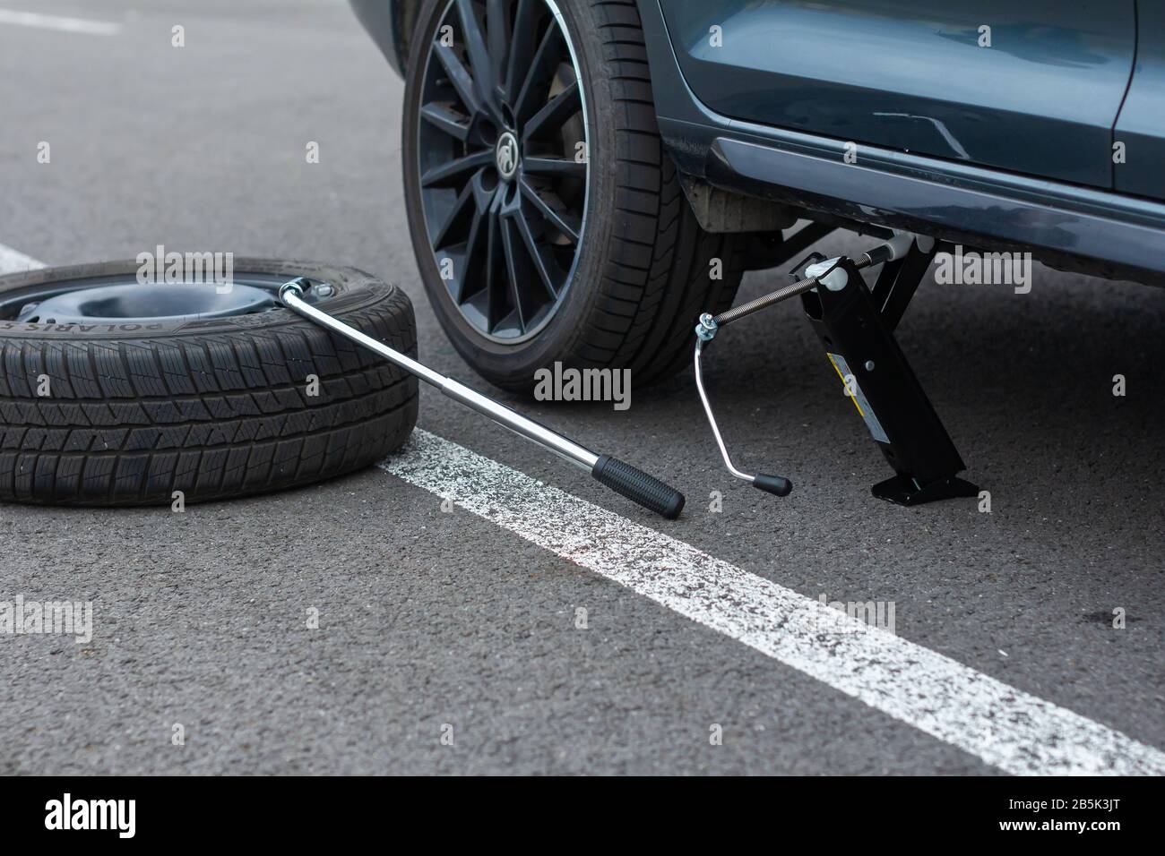 A flat tire car wheel and screwdriver are on a asphalt road on the broken car background. Jack is lifting up a vehicle. Automobile service. Tire Stock Photo
