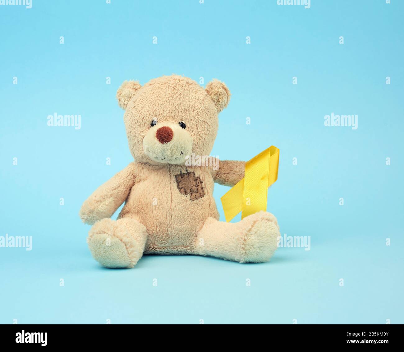 Cute brown teddy bear with a patch holds a silk yellow ribbon in the shape of a loop on a blue background, concept of the fight against childhood canc Stock Photo