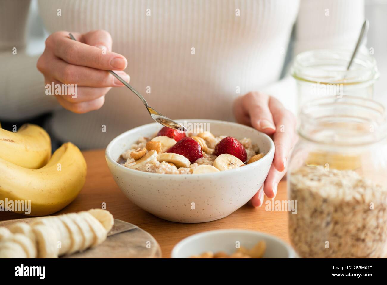 Woman eating oatmeal porridge with banana, strawberries and nuts. Healthy breakfast at the sunny morning kitchen table Stock Photo