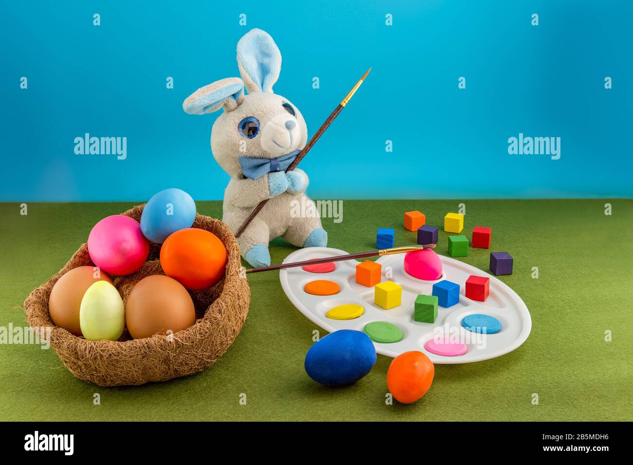 Colored easter eggs in the nest of straw. Gray plush rabbit with a palette with a brush paints Easter eggs. Multi-colored cubes around. Green grass ca Stock Photo