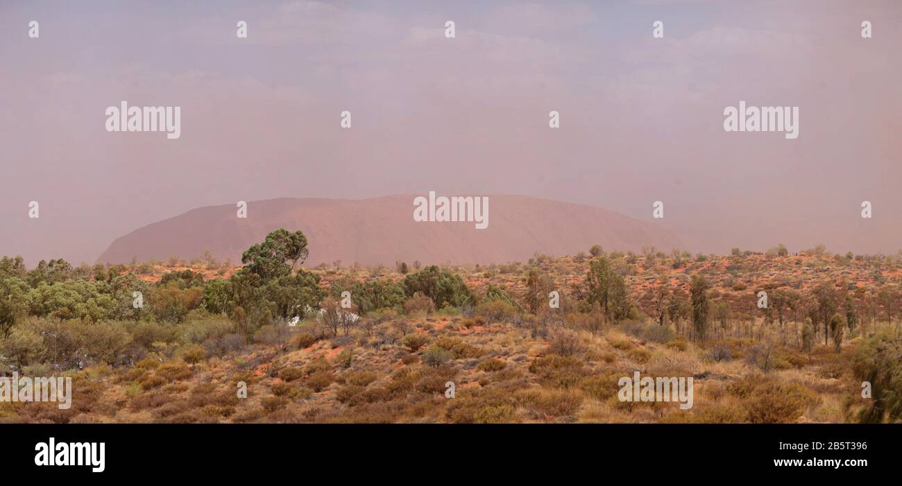 Desert dust storm blowing up a cloud of red sand obscuring the view of the massive sandstone rock of Uluru, from Imalung lookout Yulara, Australia Stock Photo