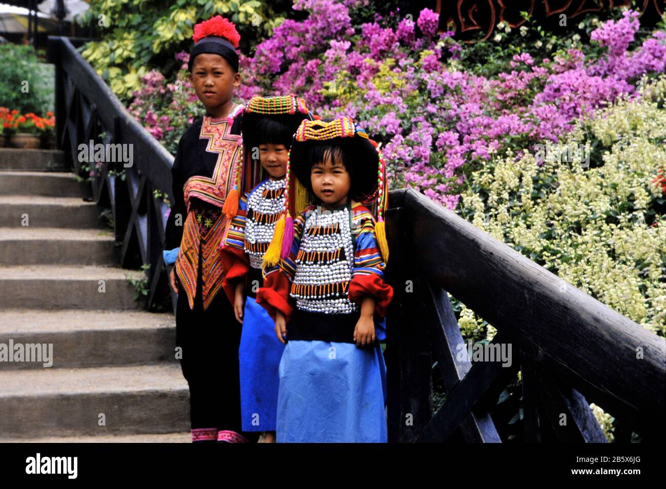 three young girls in typical colorful attire in Bangkok, Thailand in the park on the steps by bushes in full bloom Stock Photo