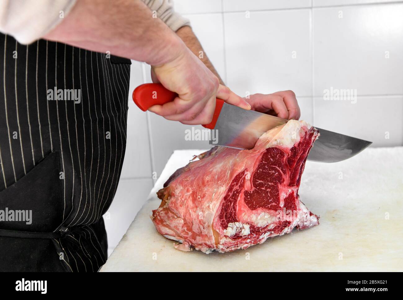 Male butcher in black apron cutting rib eye peace of ham with big knife, viewed in close-up Stock Photo