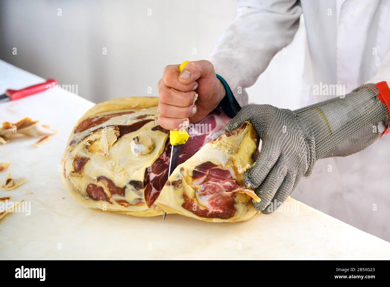 Butcher with short sharp knife deboning pice of prosciutto ham meat, using wire mesh cut resistant glove Stock Photo