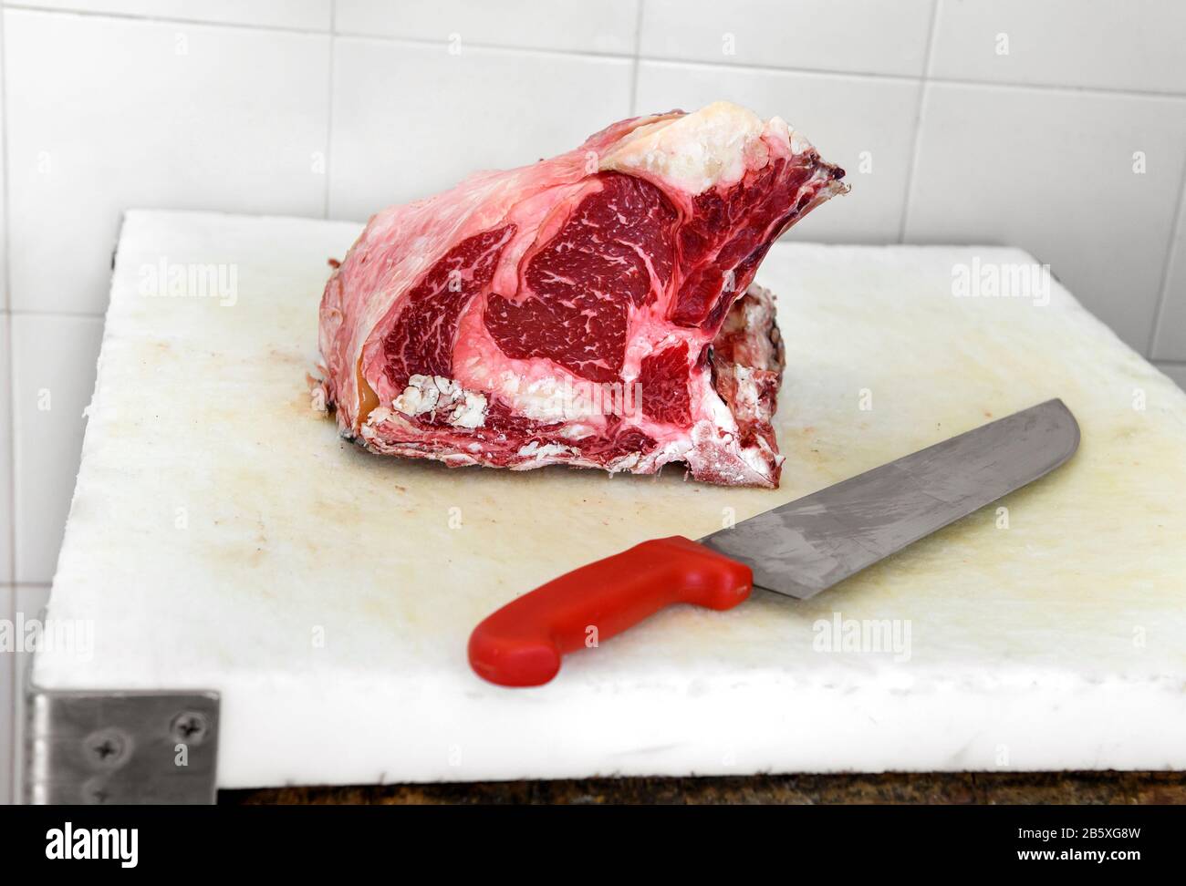 Fresh raw rib eye piece of ham meat on cutting board next to big red knife Stock Photo