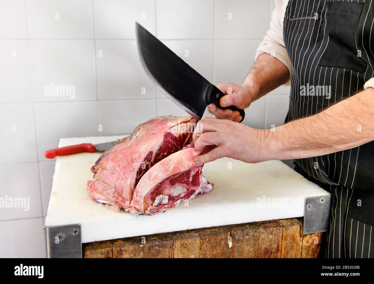 Butcher using a large cleaver to cut rib eye steaks on a butchers board in the butchery in a close up on his hands Stock Photo