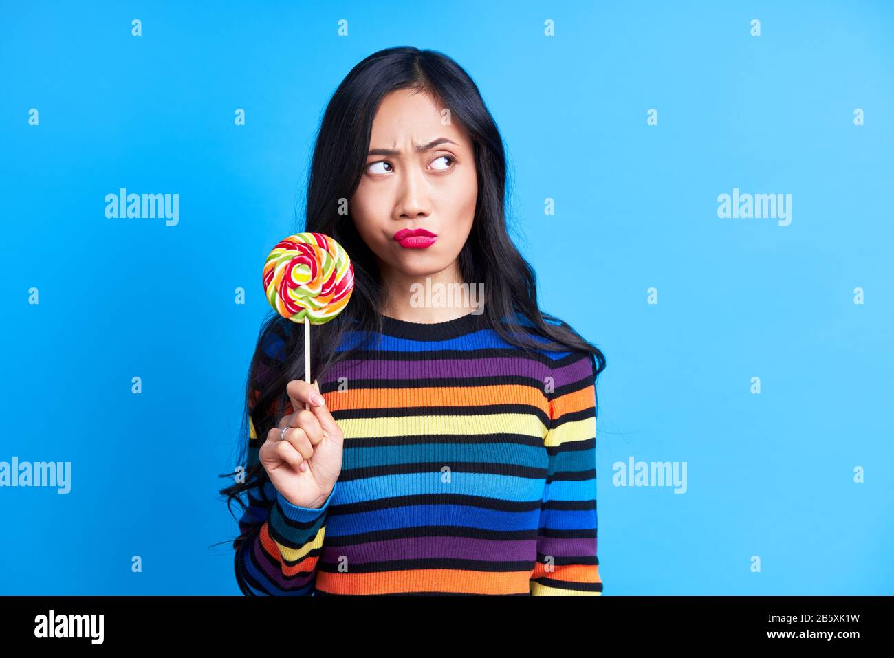 Thoughtful woman with with colorful lollipop looking away expressing doubt on blue background. Emotion concept, sweet food, diet Stock Photo
