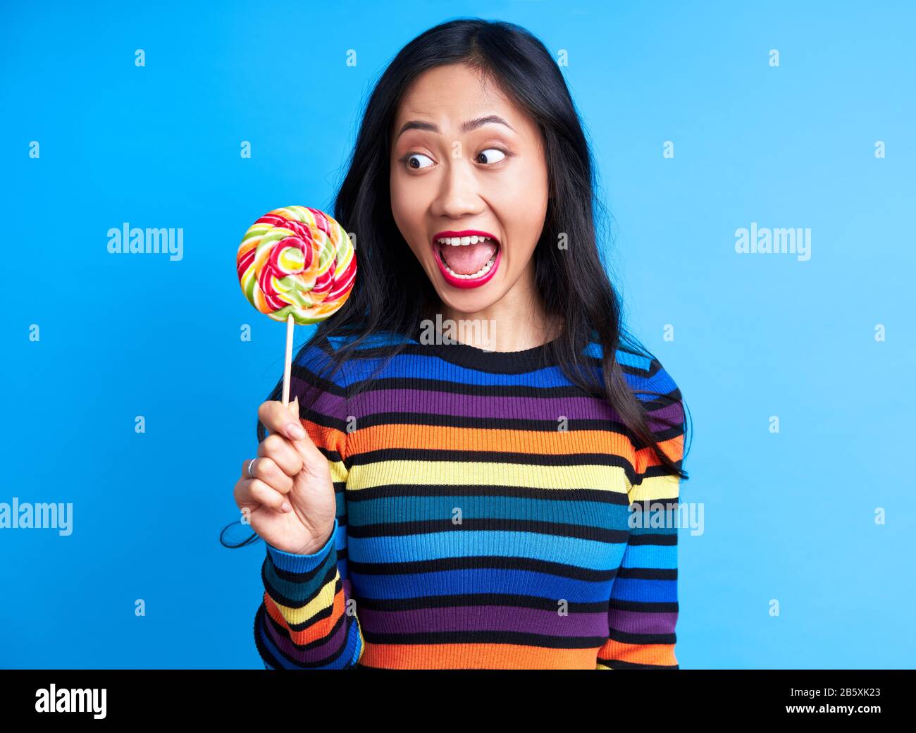 Hungry funny woman want to eat delicious colorful lollipop isolated  on blue background. Yummy, emotion concept, sweet food, diet Stock Photo