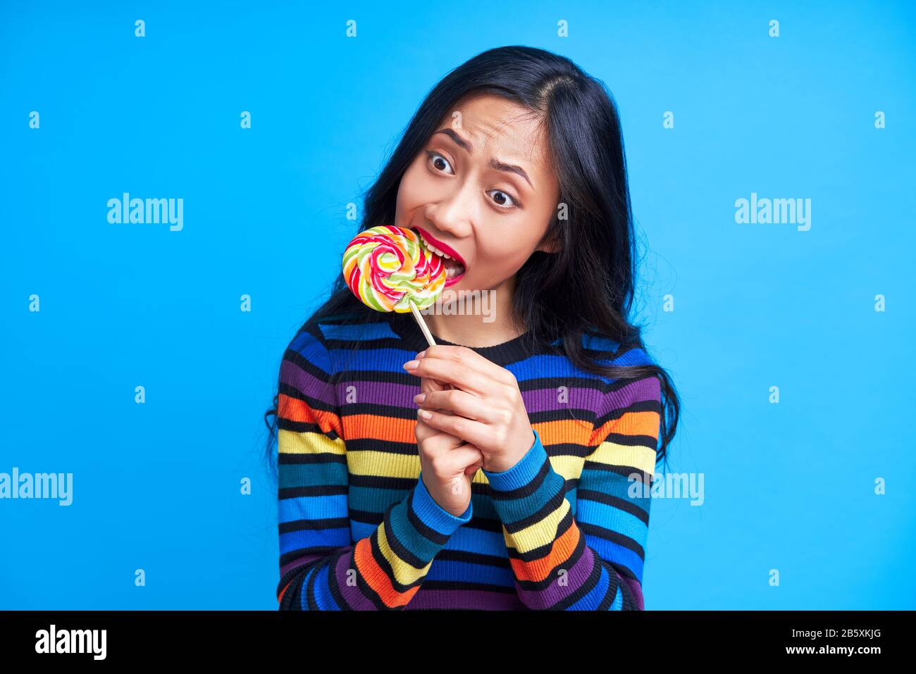 Hungry funny woman bite delicious colorful lollipop isolated  on blue background. Yummy, emotion concept, sweet food, diet Stock Photo
