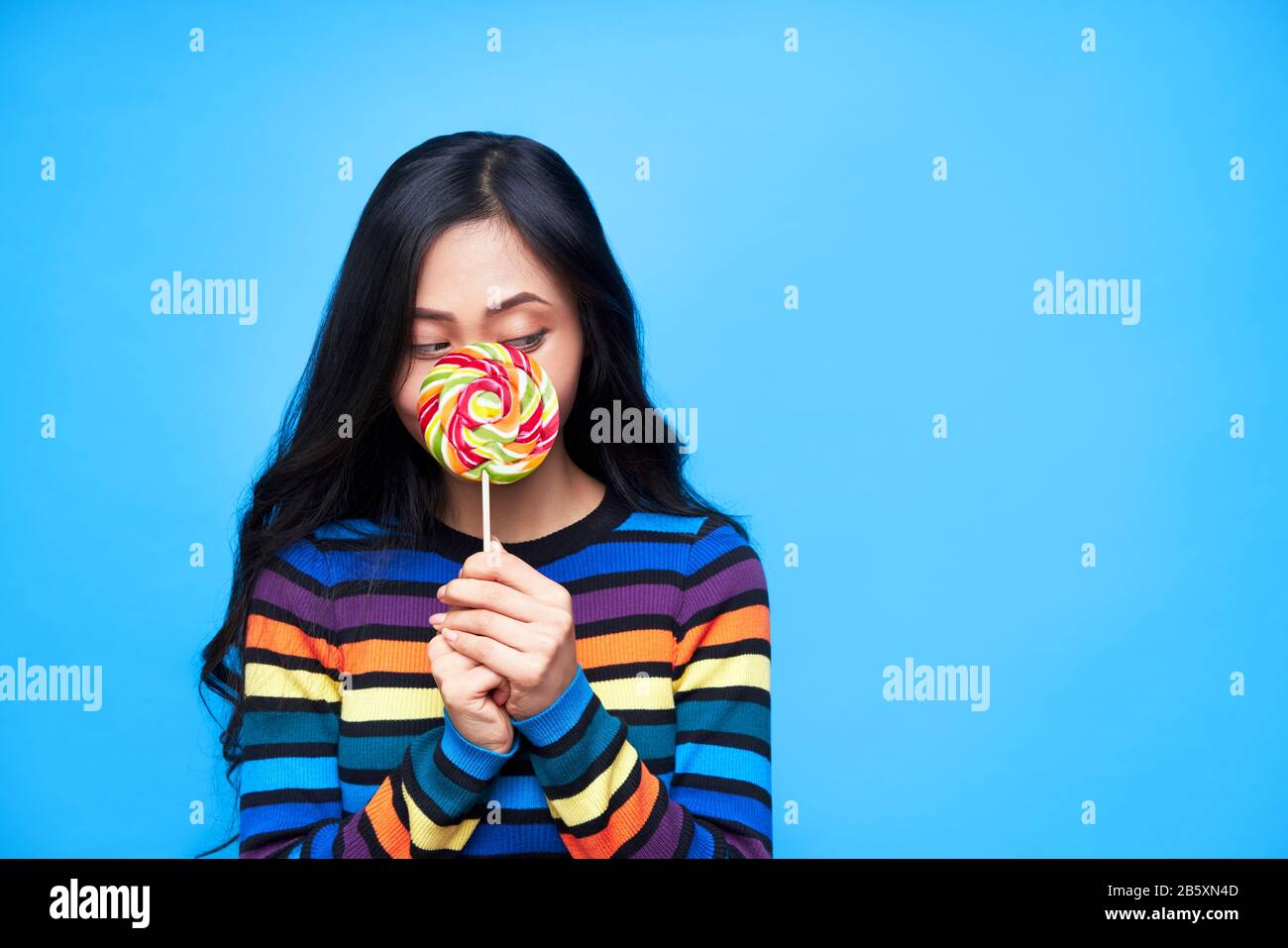 Young woman hide her face behind colorful lollipop isolated on blue background. Emotion cocept, sweet food Stock Photo