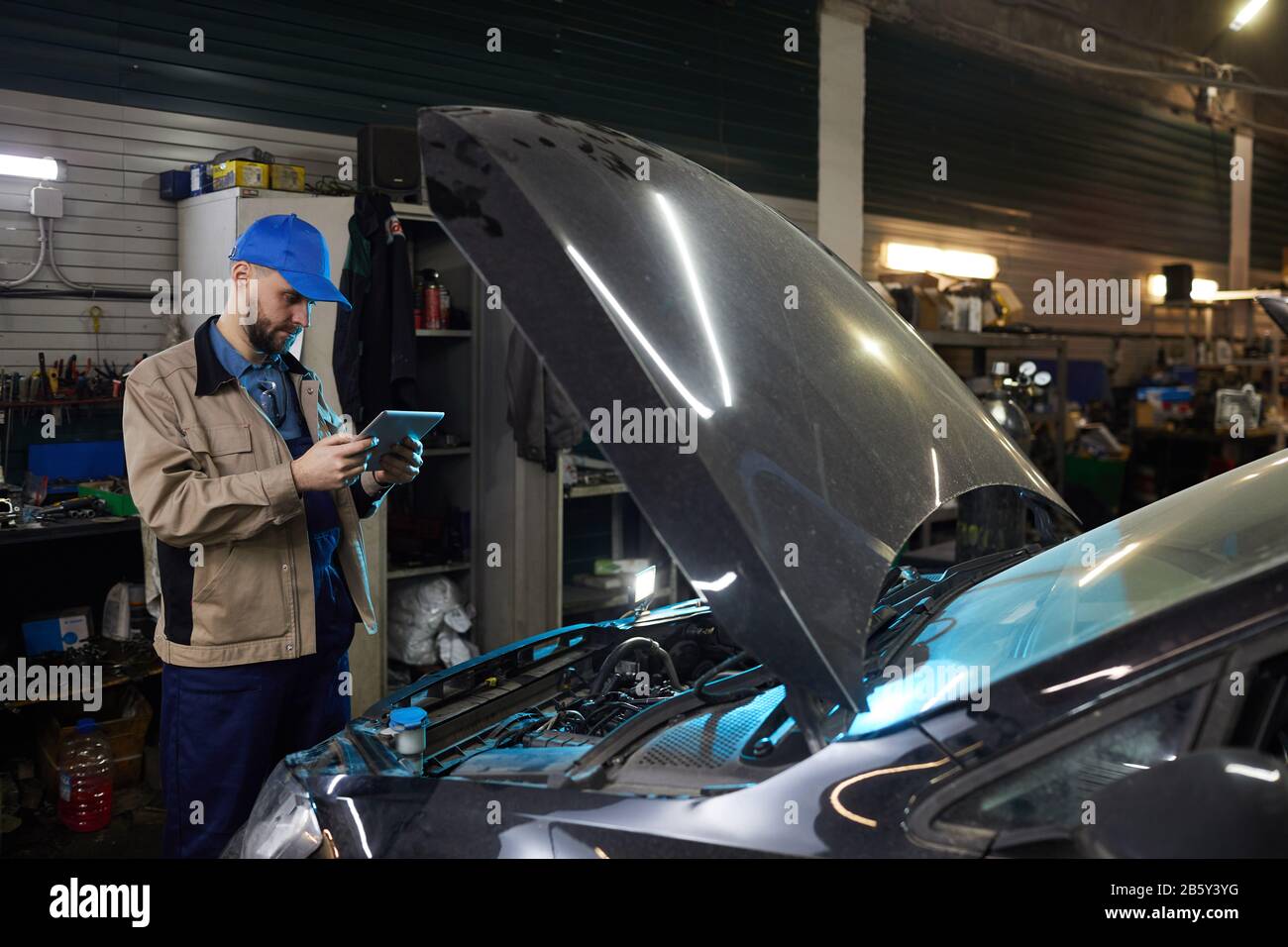Horizontal medium long shot of modern auto mechanic using tablet PC while working in auto service workshop, copy space Stock Photo