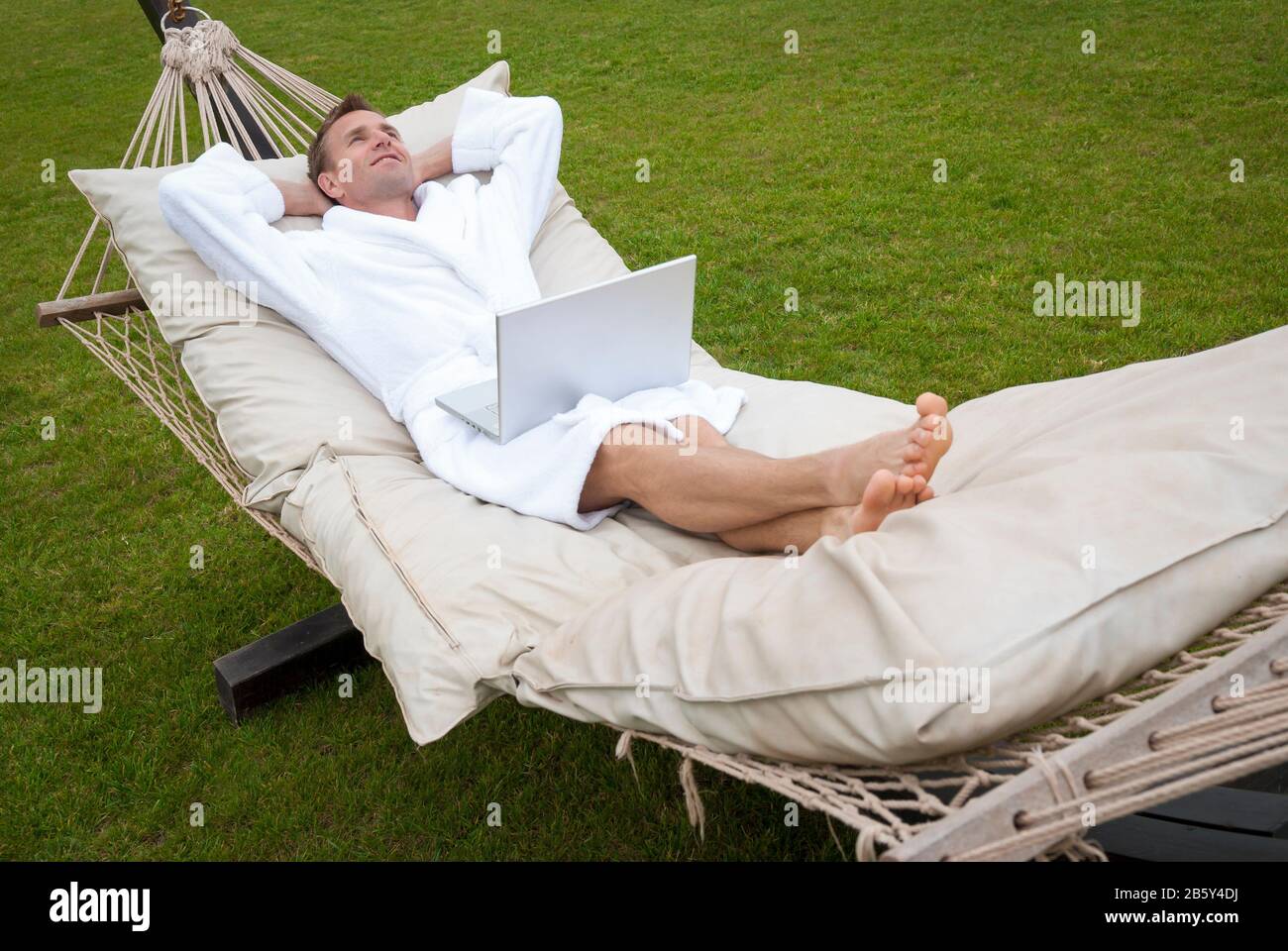 Relaxed man in plush white robe lying back with hands behind his head on a large outdoor hammock with his laptop computer Stock Photo