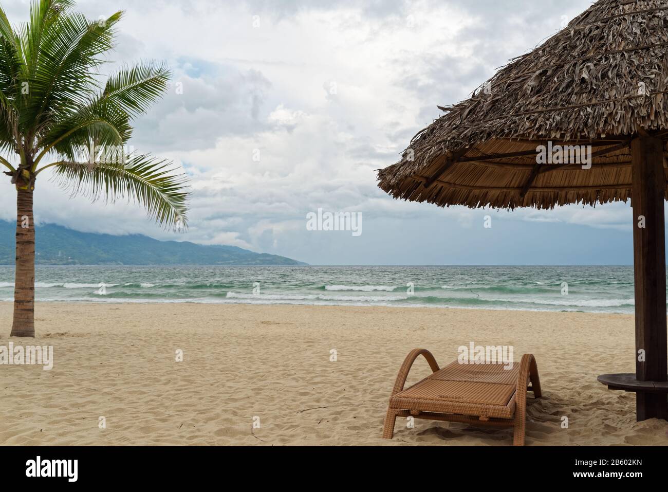 Holiday scene of lounger and straw parasol on tropical sandy beach against sea and cloudy sky. My Khe beach in Da Nang, Vietnam Stock Photo