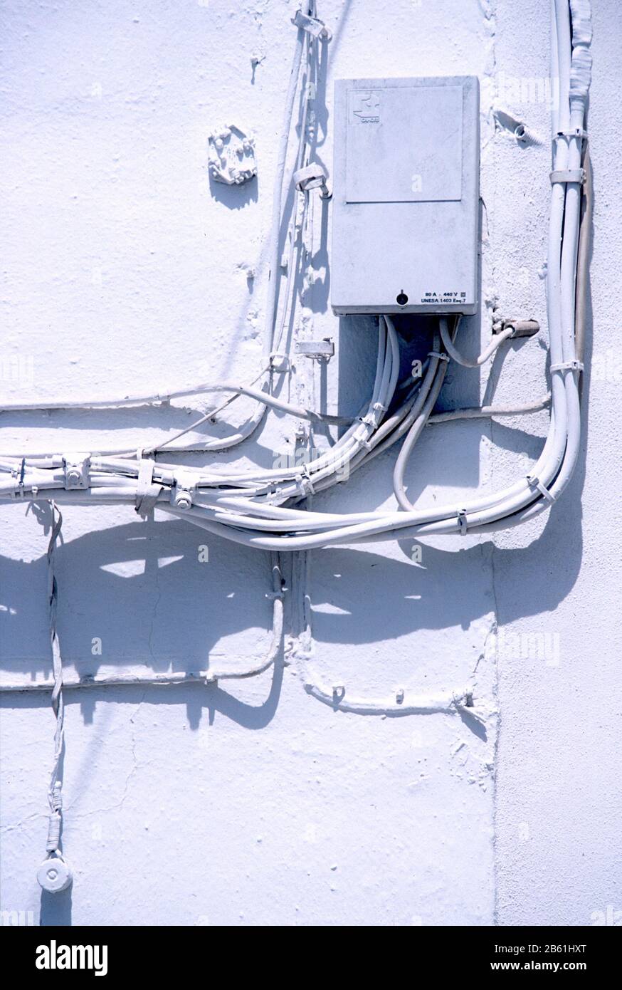 Electricity junction box on a white, painted wall in Zahara de la Sierra, Cadiz, Andalucia, Spain. Incoming wires cast shadows on the wall. Stock Photo