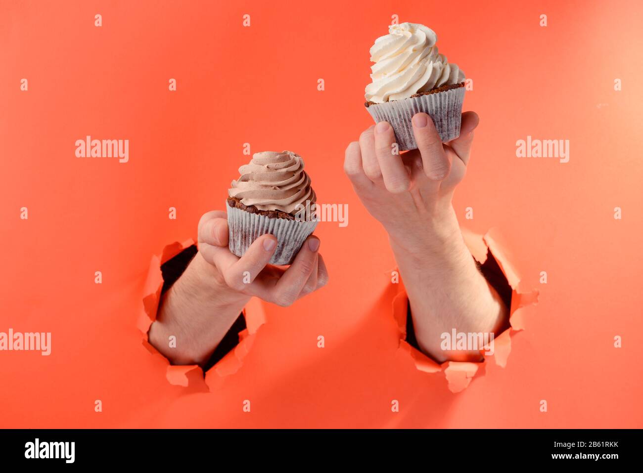 Hands holding two cupcakes through torn holes in red paper background