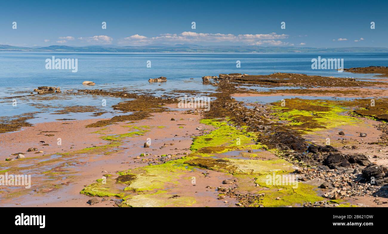 Blue seas of the Firth of Clyde lap at the rocky shore of the Isle of Arran on the west coast of Scotland, with the hills of Ayrshire in the distance. Stock Photo