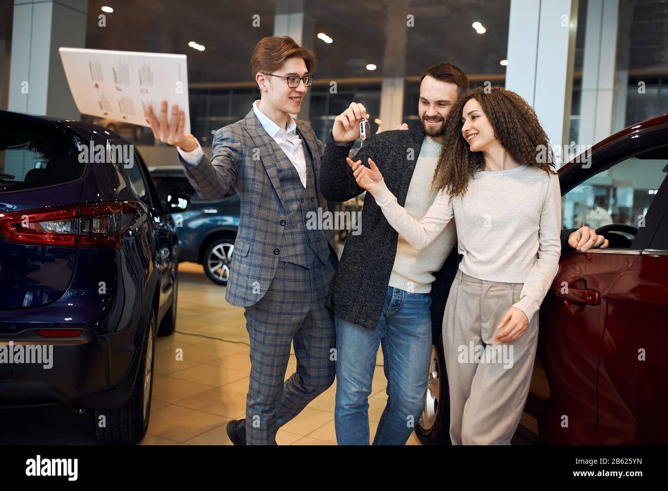 successful professional cheeful dealer is handing auto keys to a young couple, they are excited, cars standing in the background, bargain, dream comes Stock Photo