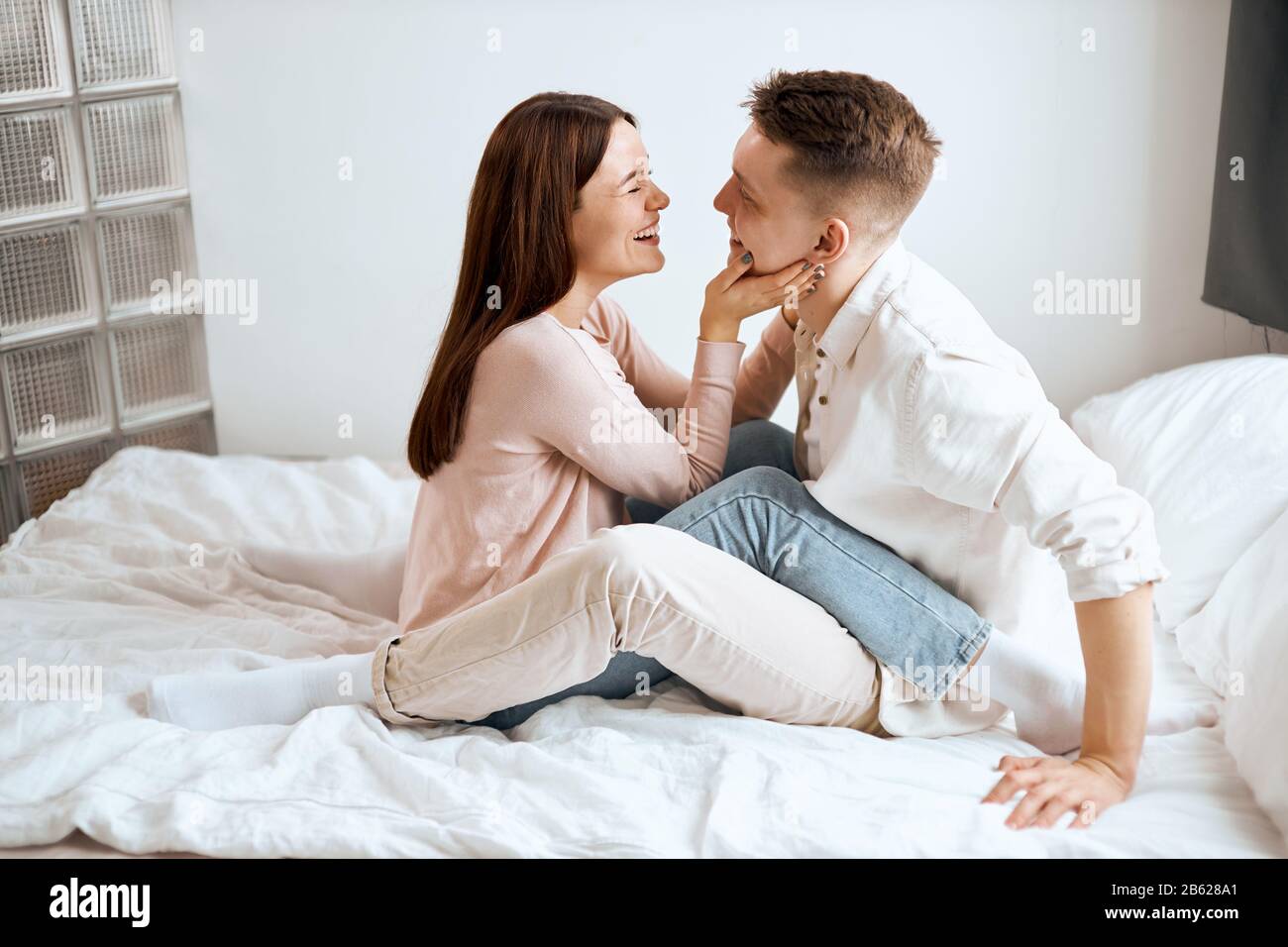 young attractive man and woman relaxing, embrace and have fun sitting on the bed.side view full length photo, couple laughing in the bedroom, man tell Stock Photo