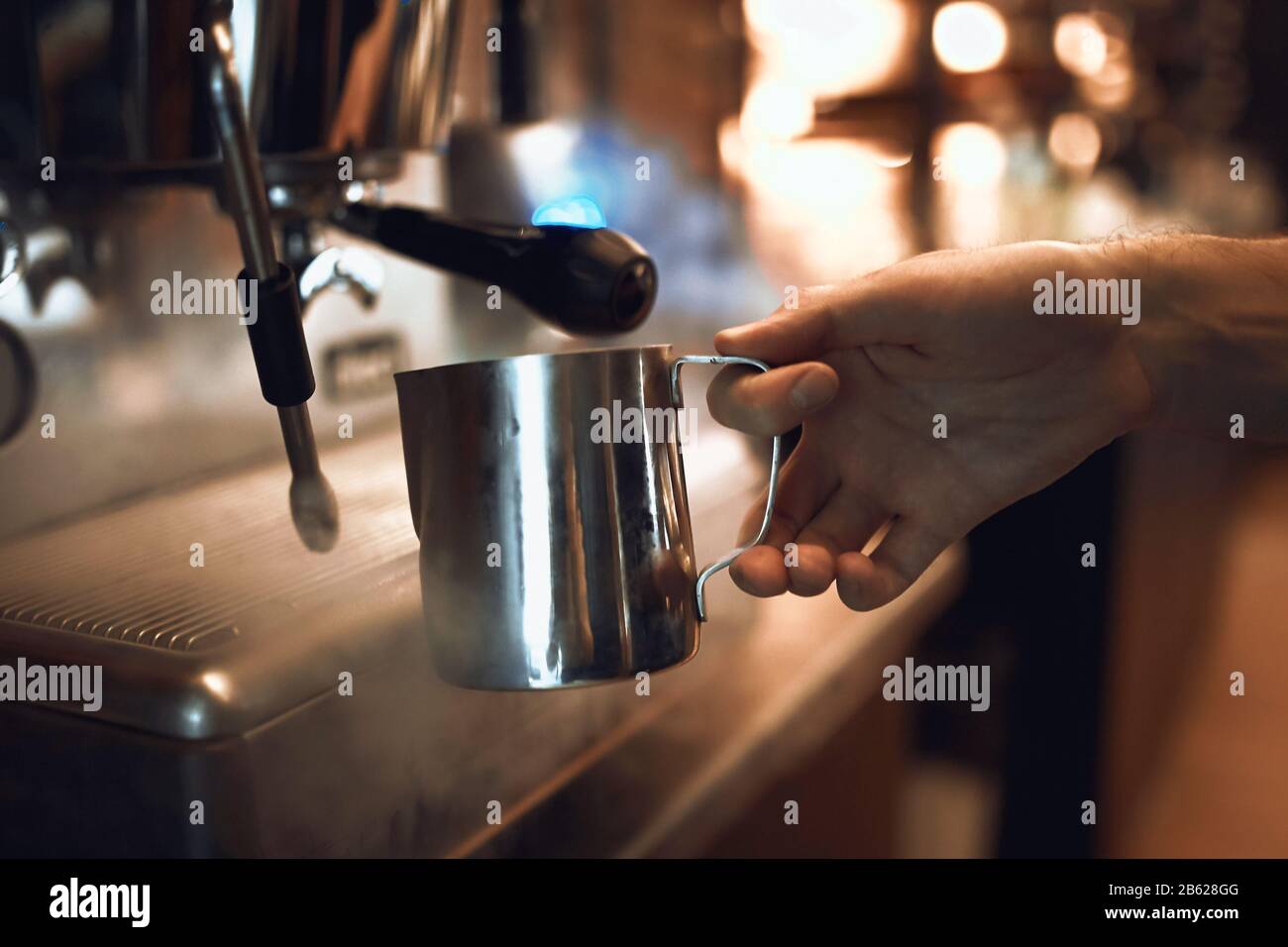 professional barista is holding a cup of milk, standing near a coffee machine, close up side view photo. Stock Photo