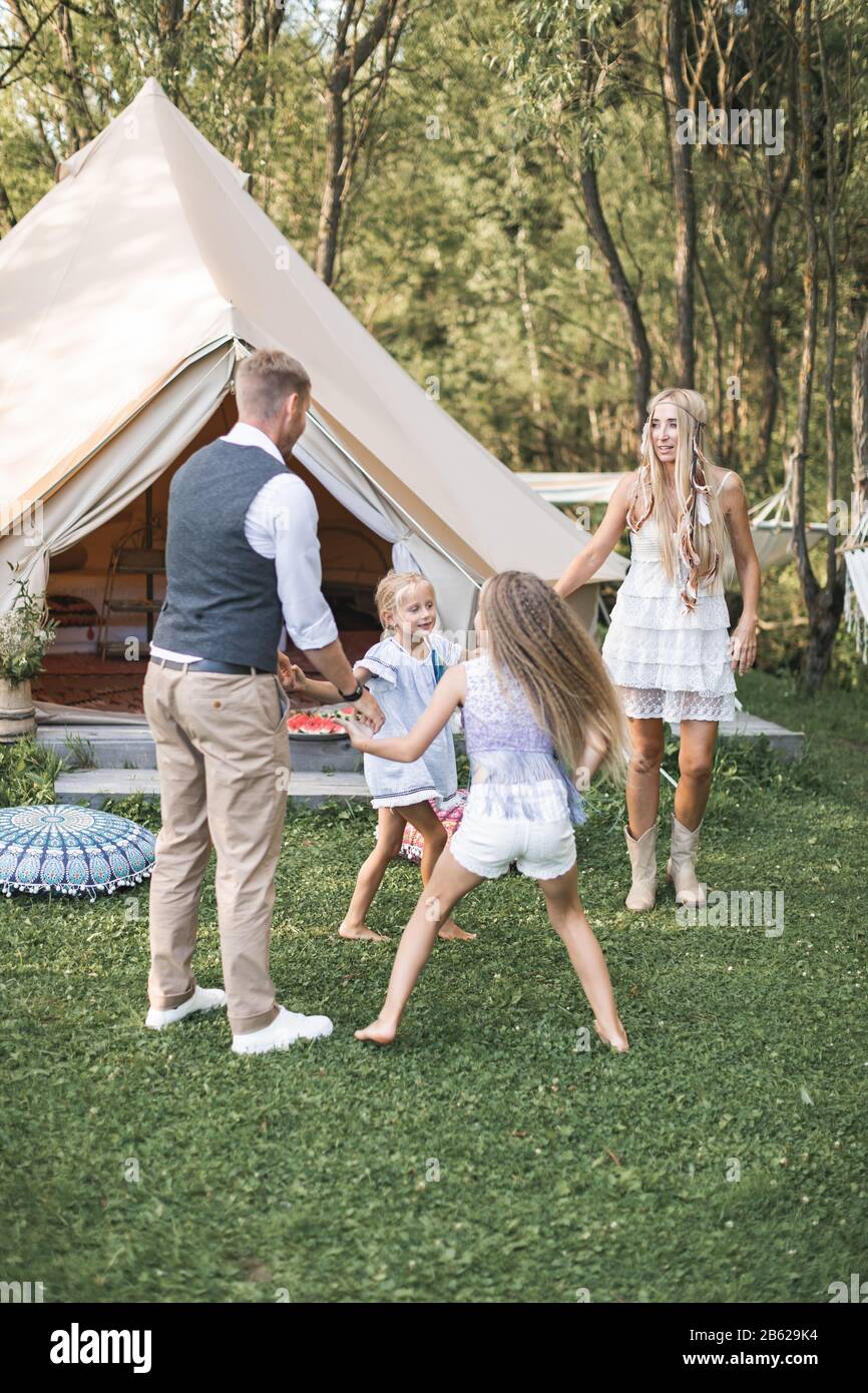 Young family is playing together on a sunny day in park. Father, mother and two daughters in casual boho clothes dancing and jumping, holding hands in Stock Photo