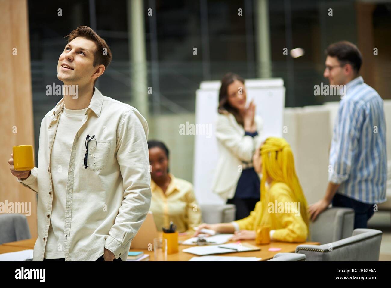 cheerful pleasant guy with cup of tea lookingup, dreaming to have holidaym, his friends talking in the blurred background, studio shot. love, man thin Stock Photo