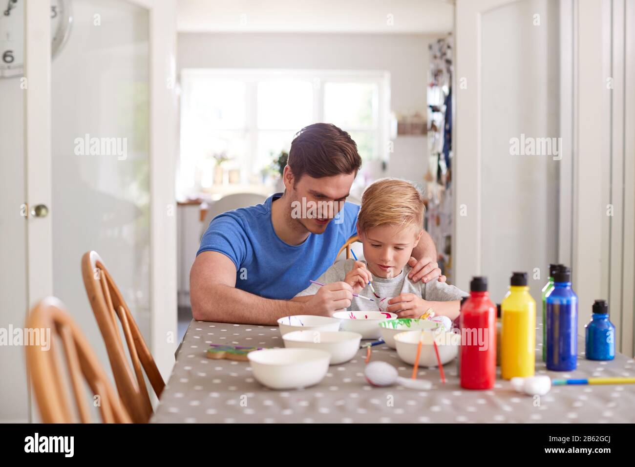 Father With Son Sitting At Table Decorating Eggs For Easter At Home Stock Photo