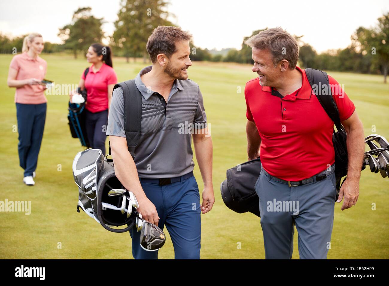 Two Mature Couples Playing Round Of Golf Carrying Golf Bags Along Fairway Stock Photo