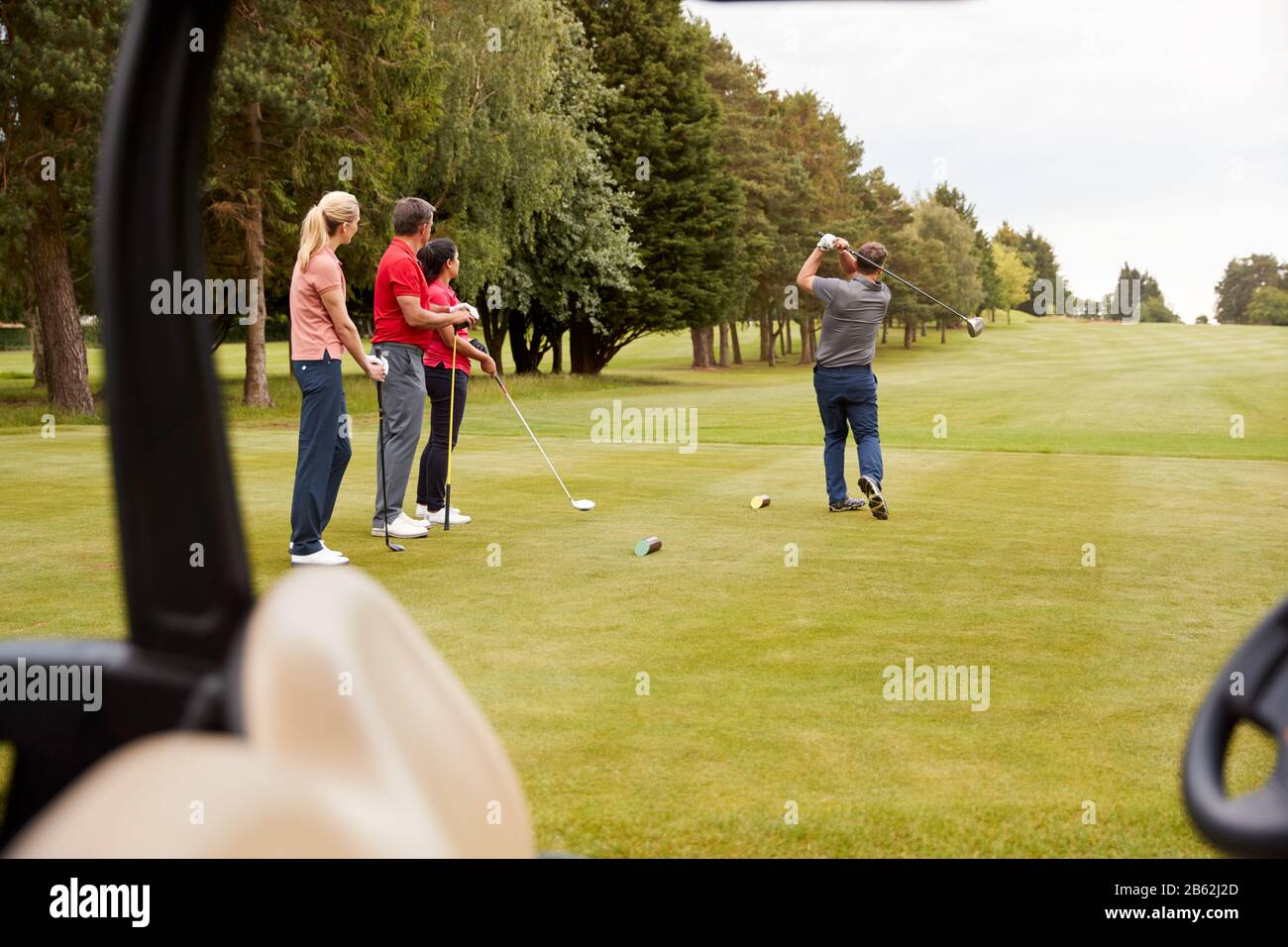 Two Couples Golfing Hitting Tee Shot Along Fairway With Driver With Buggy In Foreground Stock Photo