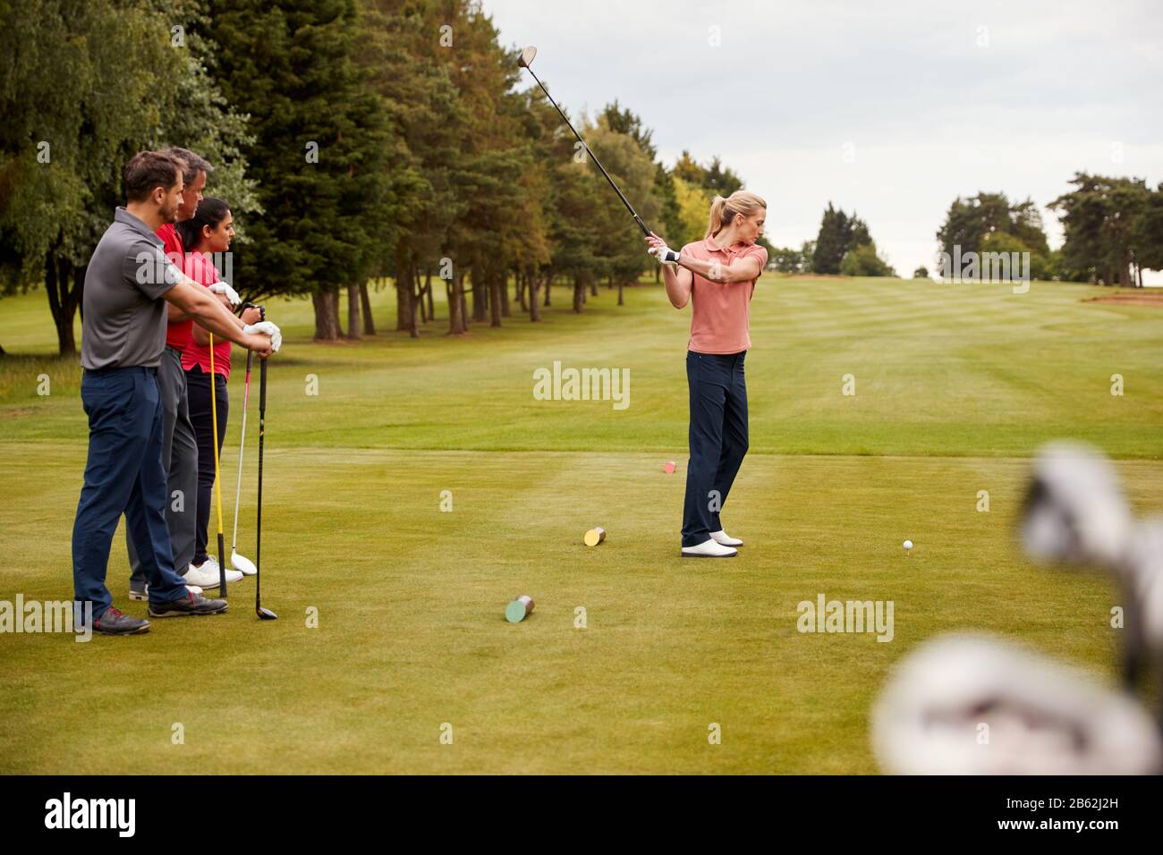 Two Couples Golfing Hitting Tee Shot Along Fairway With Driver With Clubs In Foreground Stock Photo