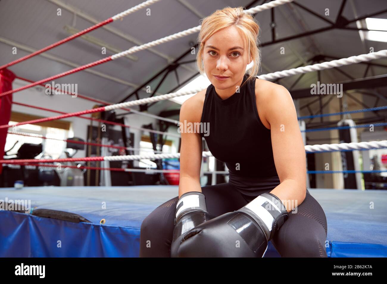 Portrait Of Female Boxer With Gum Shield In Gym Wearing Boxing Gloves Sitting On Boxing Ring Stock Photo