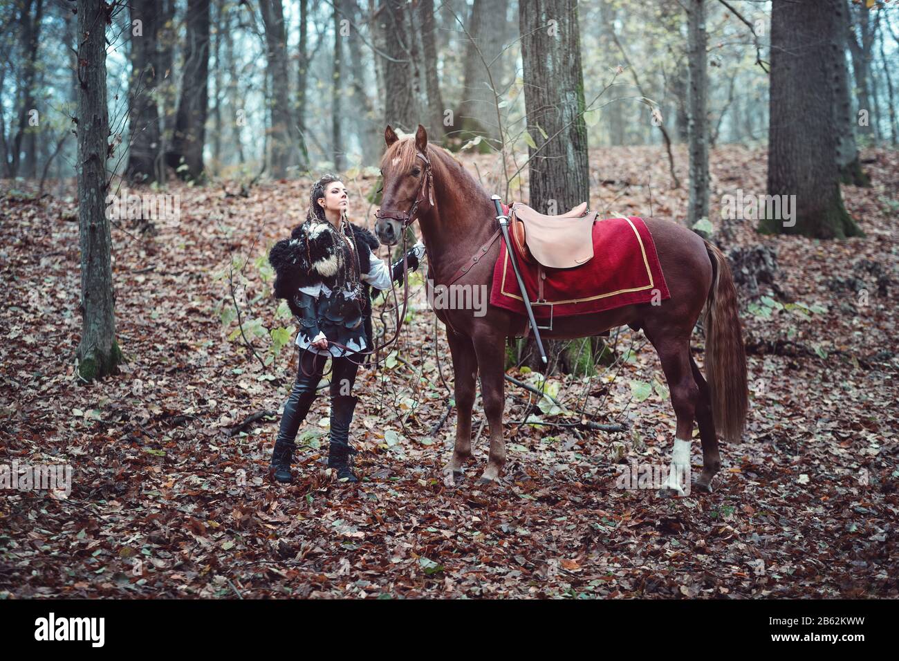 Warrior woman with her brown horse into the woods - Beautiful scandinavian viking girl - Medieval cinematic movie scene Stock Photo