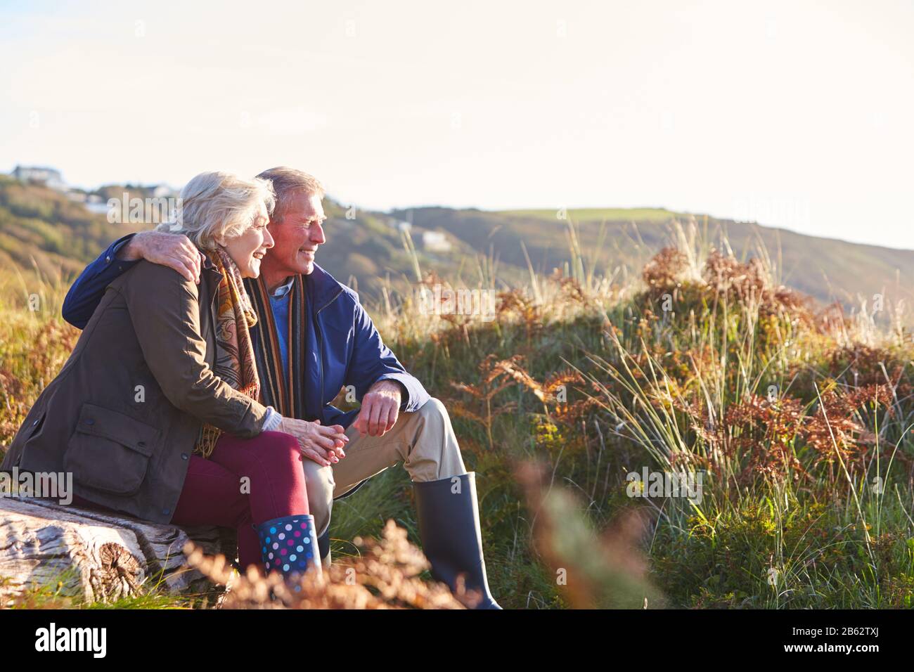 Loving Active Senior Couple Walking Along Coastal Path In Autumn Resting On Rock Together Stock Photo