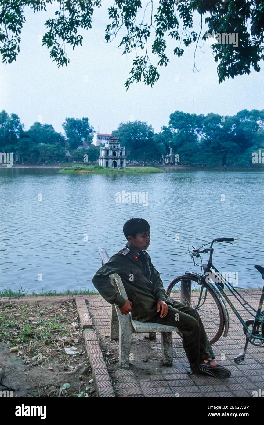 Soldier seated by Hoan Kiem Lake, also known as Sword Lake, with the Turtle Tower behind, Hanoi, Vietnam, November 1995 Stock Photo