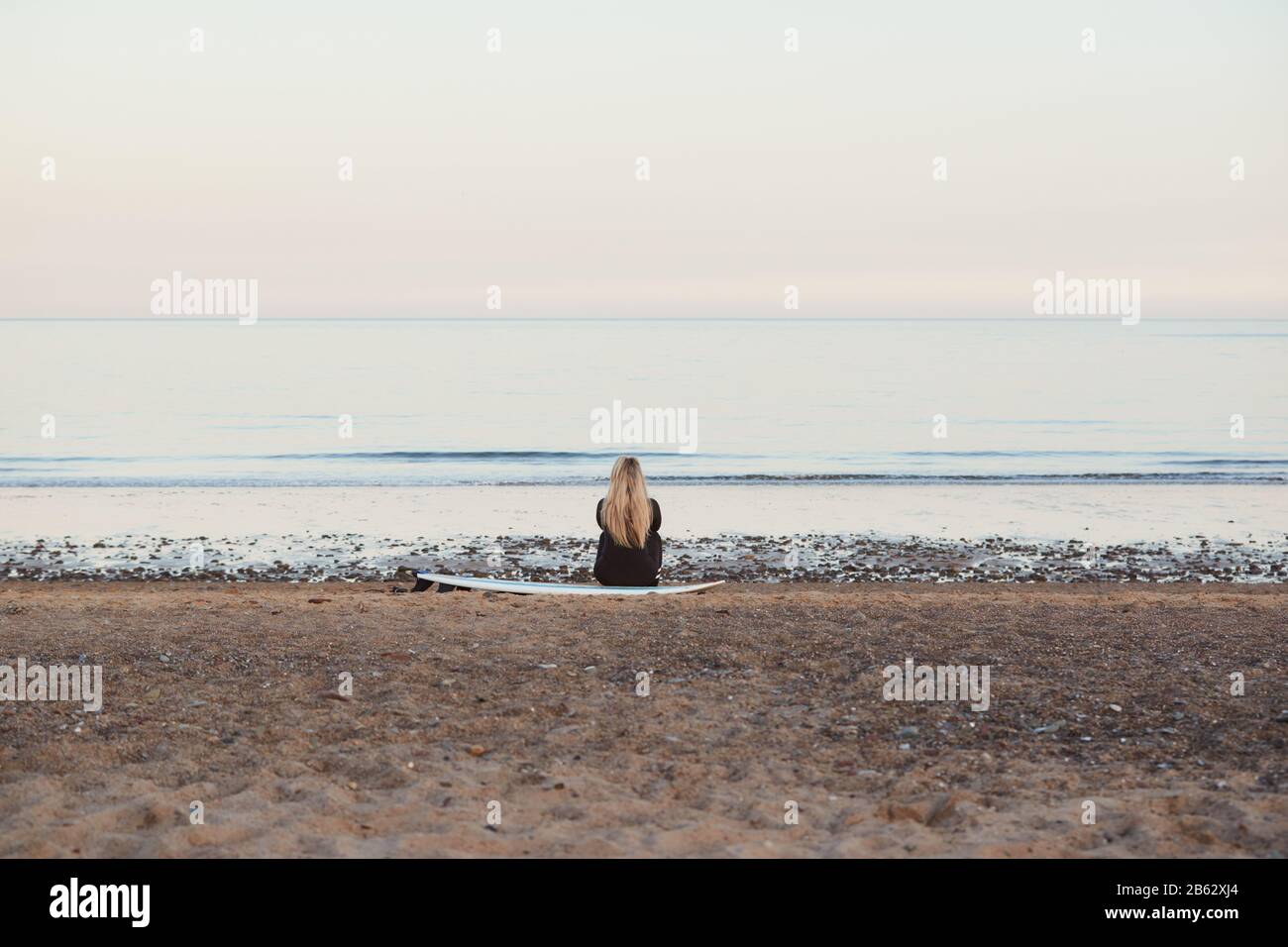 Rear View Of Thoughtful Woman Wearing Wetsuit On Surfing Staycation Looking Out To  Sea At Waves Stock Photo