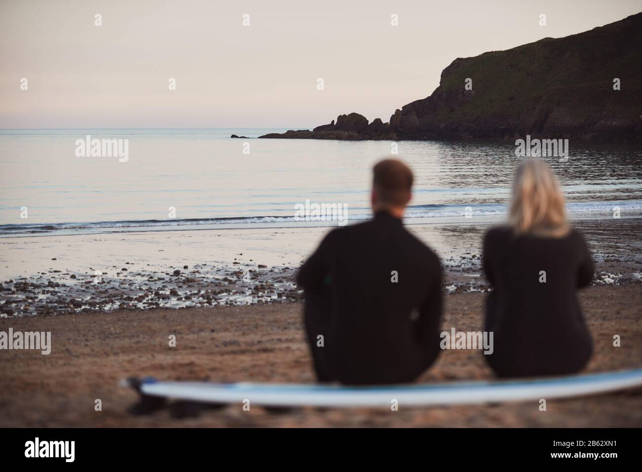 Defocused Shot Of Couple In Wetsuits On Surfing Staycation Sitting On Surfboard Looking Out To  Sea Stock Photo