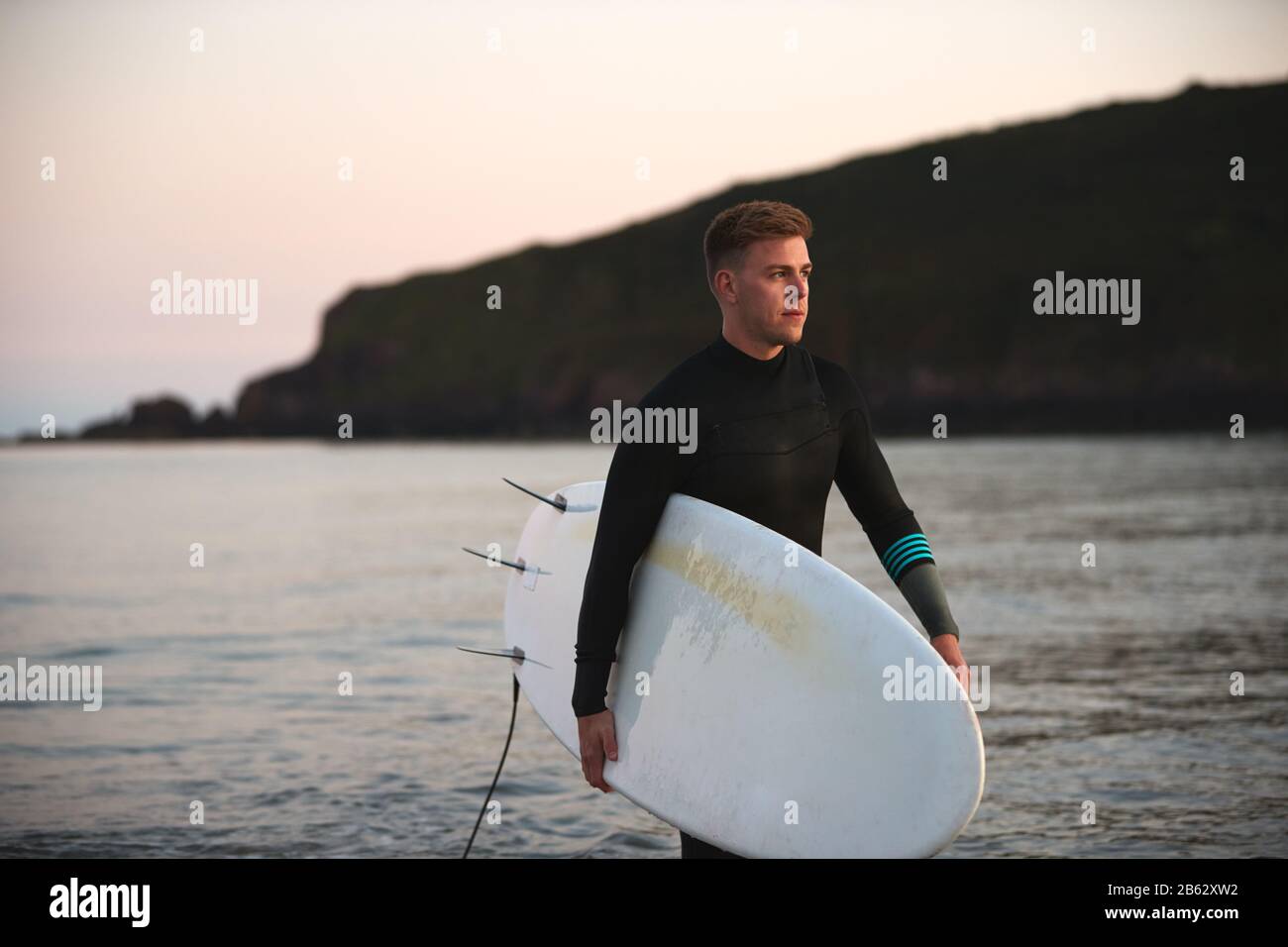 Man Wearing Wetsuit Carrying Surfboard As He Walks Out Of Sea Stock Photo