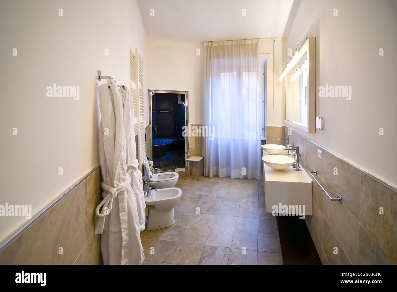 A large bathroom with double basin sinks, bidet, hanging robes, toilet and large walkin shower