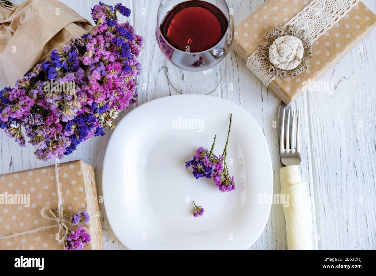 Table setting. Plate on wooden background with gift box, cutlery, a glass of red wine and flowers. Stock Photo