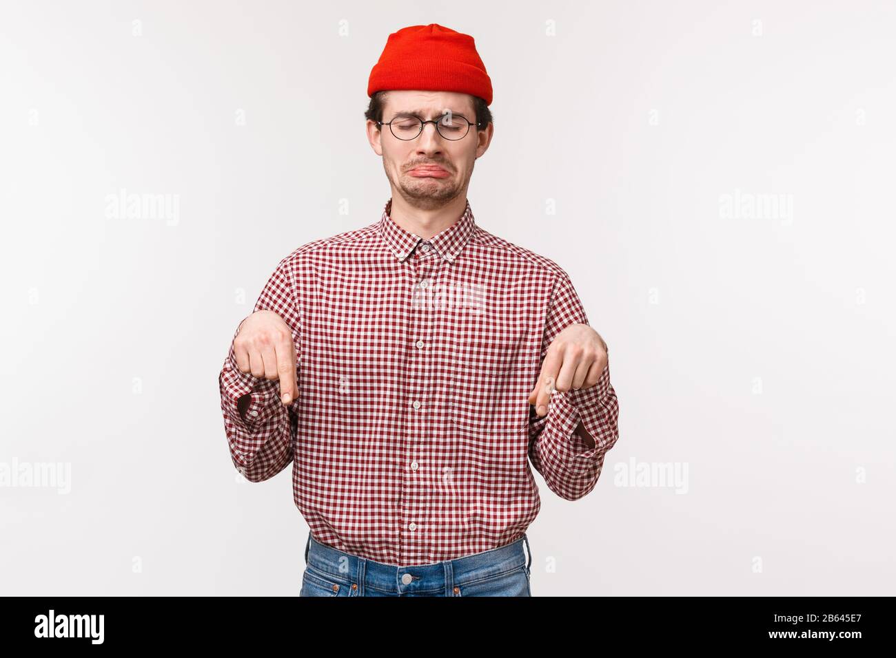 Waist-up portrait distressed and upset whining young upset guy in red beanie, sobbing and grimacing sad as pointing looking down with regret or Stock Photo