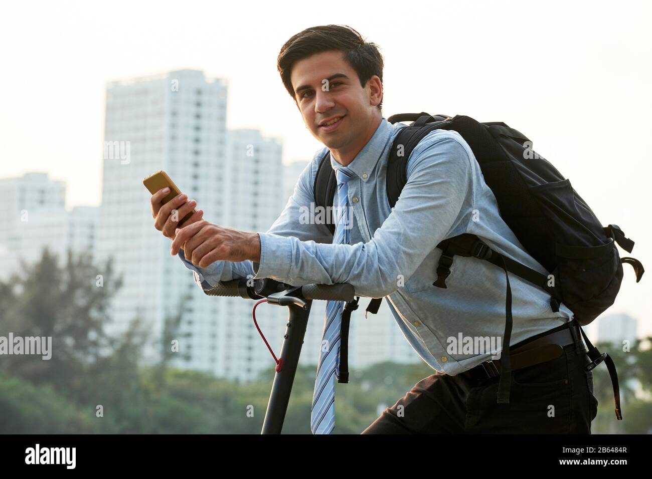Portrait of handsome young smiling businessman with big backpack holding smartphone and riding on electric scooter Stock Photo