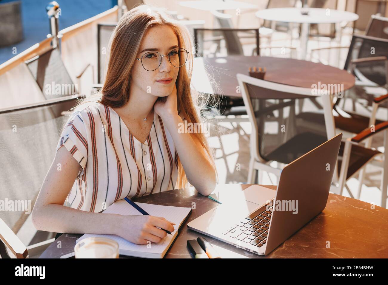 Caucasian woman with red hair and freckles having a coffee break in a coffee shop while using a laptop and making some notes Stock Photo