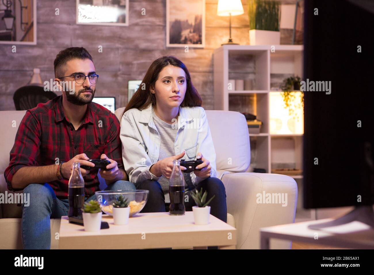 Beautiful young couple having fun playing video games with controllers at night. Couple sitting on sofa. Stock Photo