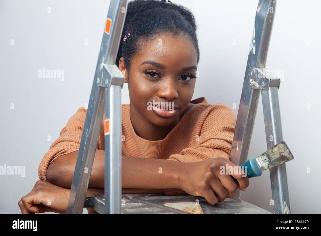 A beautiful young woman of African ethnicity resting her hands on a step ladder and holding a wallpaper scraper in her hand Stock Photo