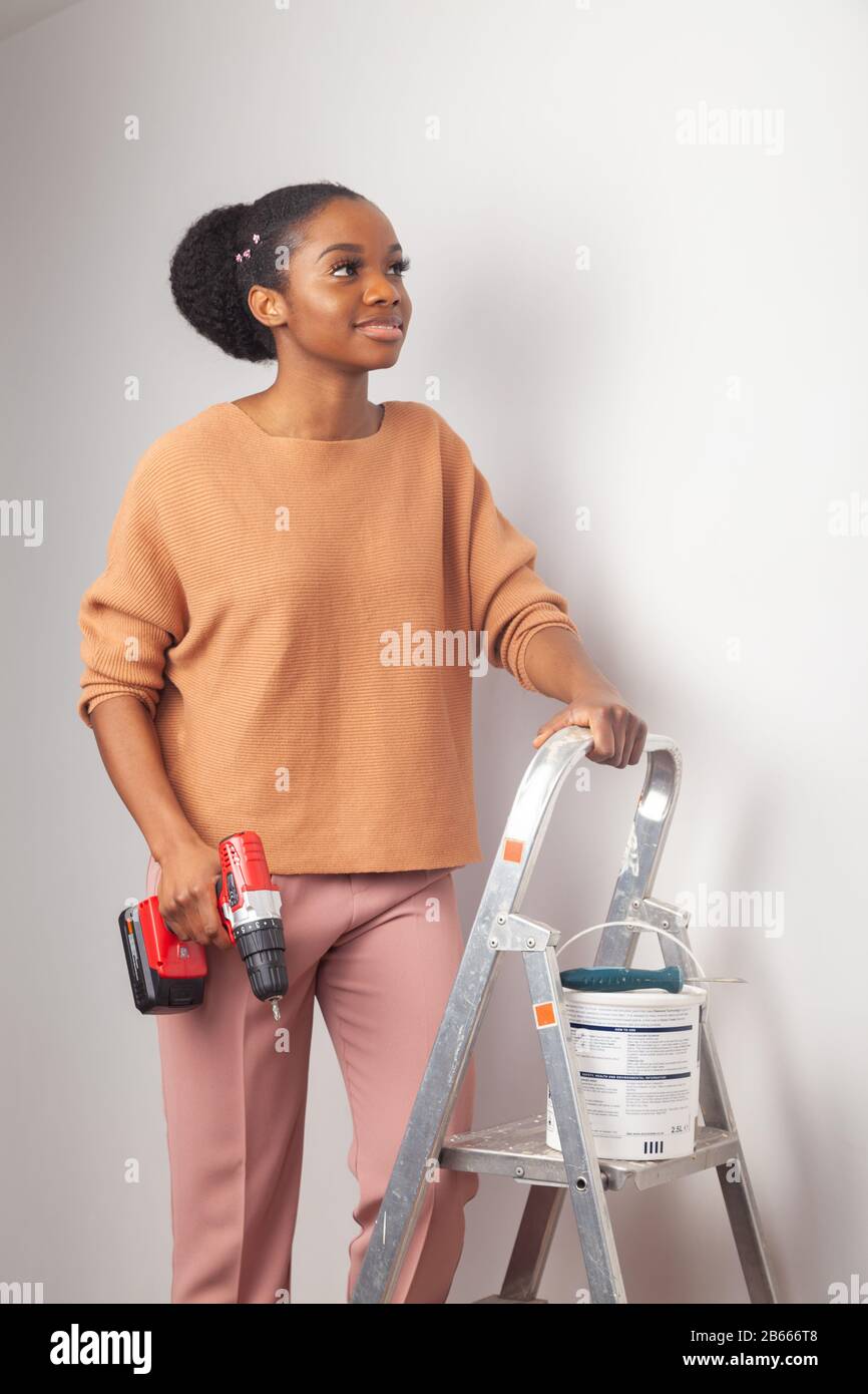 A beautiful young woman of African ethnicity standing next to a step ladder and holding an electric drill Stock Photo